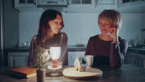 la hija se sienta con la madre, compartiendo un momento afectuoso mientras admiran la casa de pasteles bellamente decorada en una bandeja de madera, la cálida luz de las velas y la planta en olla en el fondo