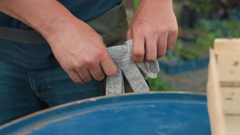 gardener wearing pocket apron picks up work gloves, tucks pair into side pocket beside blue metal drum and wooden crate, close view of hands preparing for garden task