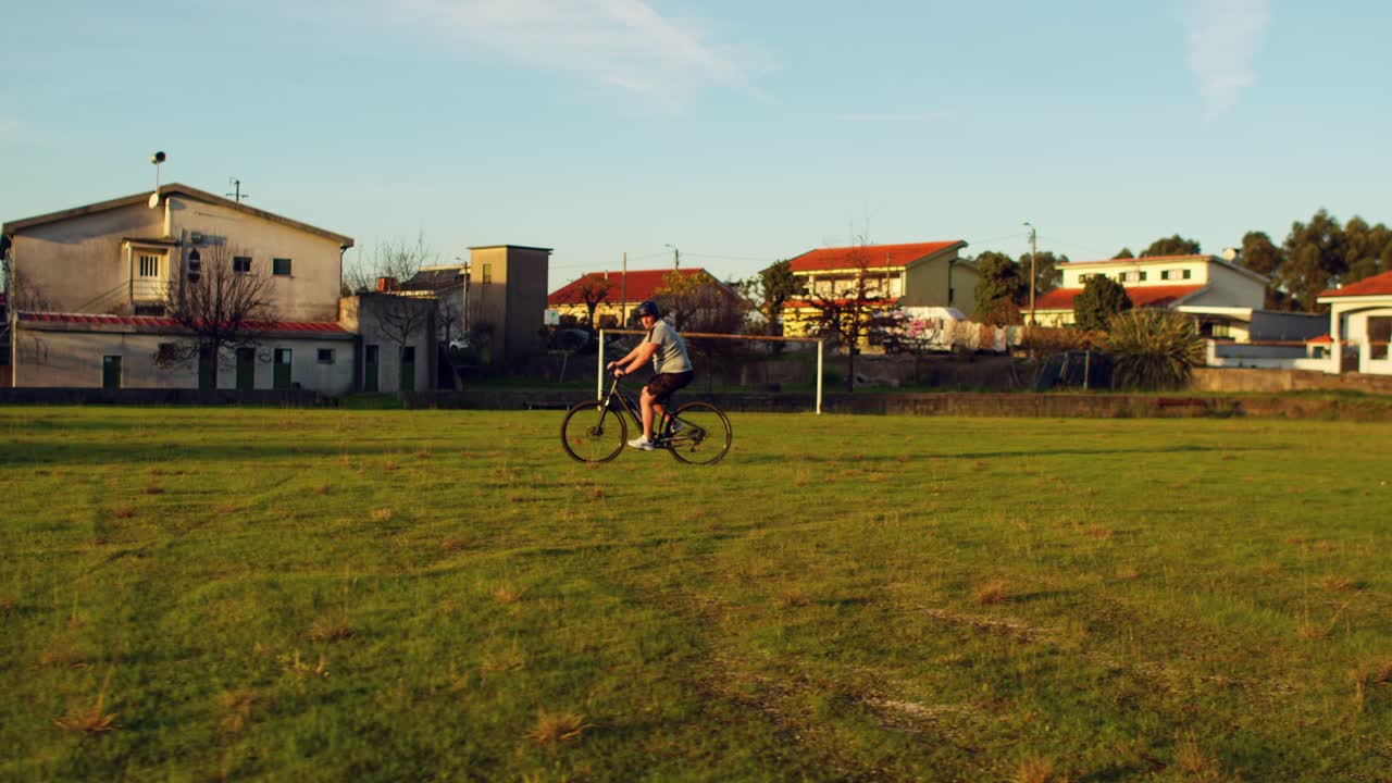 Um homem vira sua bicicleta e pedala em direção à câmera em um campo rural ao pôr do sol.