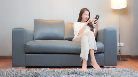 While-sitting-on-a-comfortable-grey-leather-couch-a-pretty-young-woman-focuses-on-her-smartphone-as-she-inputs-her-next-text