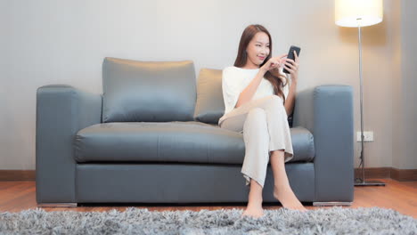 편안한 회색 가죽 소파에 앉아 예쁜 젊은 여성이 다음 텍스트를 입력하면서 스마트폰에 집중하고 있습니다.