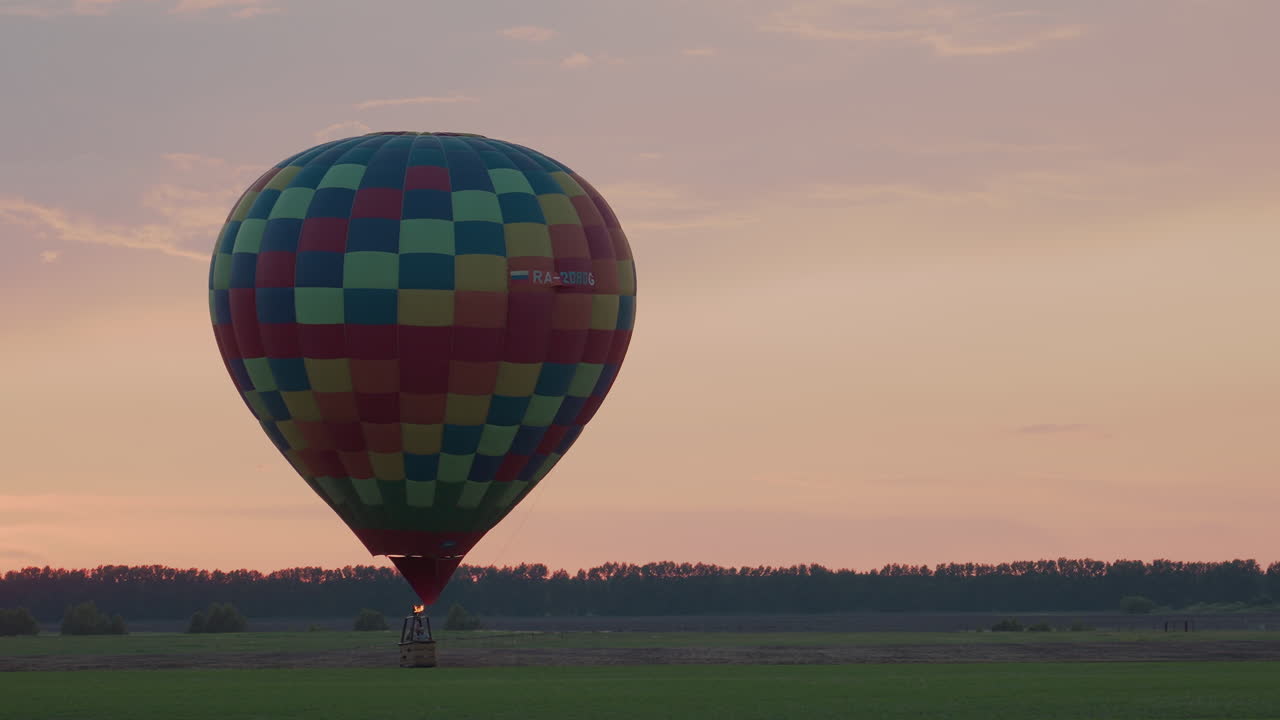 wide sunset field with colorful hot air balloon soaring in distance as burner flame bursts skyward against pastel horizon capturing peaceful evening flight adventure serene moment