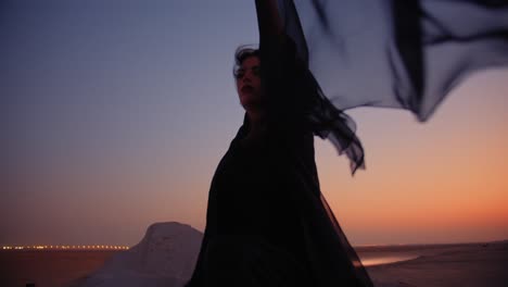 A-girl-in-a-black-abaya-sitting-on-a-fossil-rock-in-the-desert-while-the-sunset-wind-blowing-her-scarf-medium-shot-fossil-dunes