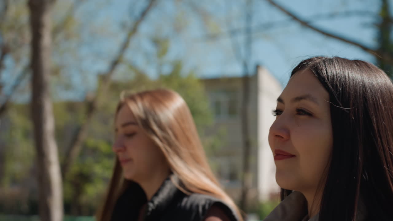 공원을 거닐며 미소 짓는 백인과 아시아인의 클로즈업 프로필, 친구 간의 솔직한 채팅, 햇빛이 비치는 나무와 얕은 피사계 심도, 자연스러운 웃음과 편안한 친밀감, 젊은 크리에이터