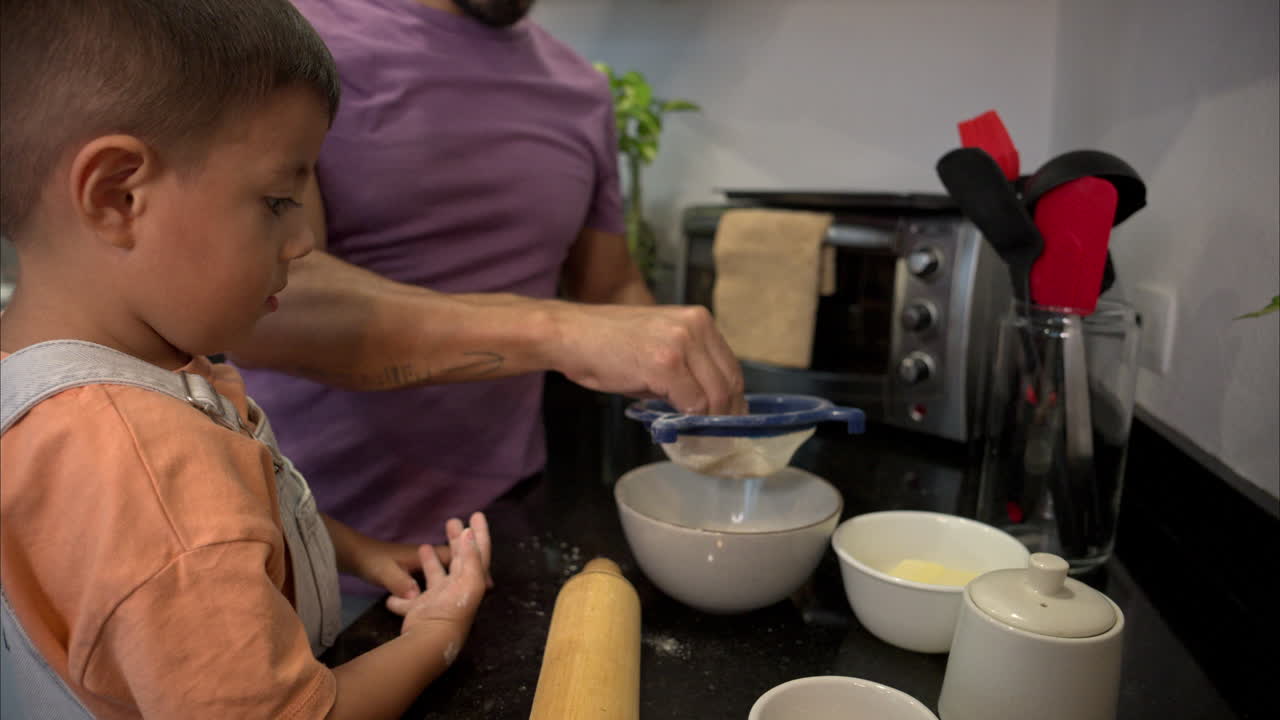 Cámara lenta de un niño latino mexicano pequeño viendo a su padre tamizar harina con sus manos en un colador de plástico en la cocina.