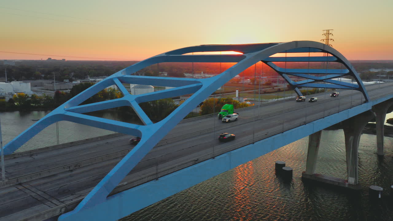 A blue steel arch bridge rises gracefully over calm river water, illuminated by the warm orange light of sunset that glows between its sweeping geometric curves