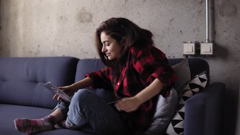 una hermosa chica de unos 20 años escribiendo algo en el teclado de una computadora portátil mientras se relaja en el sofá