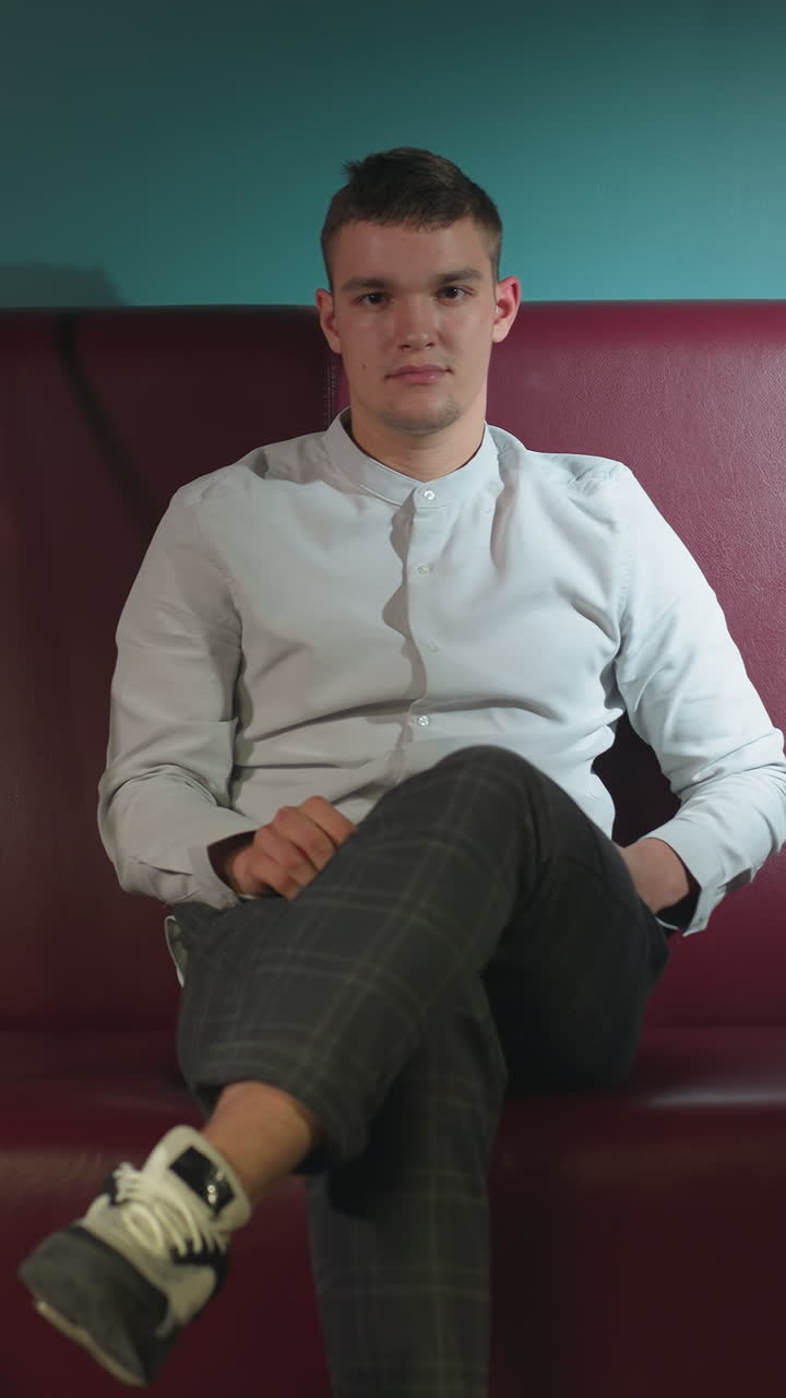 un hombre con camisa blanca y pantalones a cuadros se sienta en un sofá rojo, rodando una pelota de billar naranja en la mano con una expresión seria.