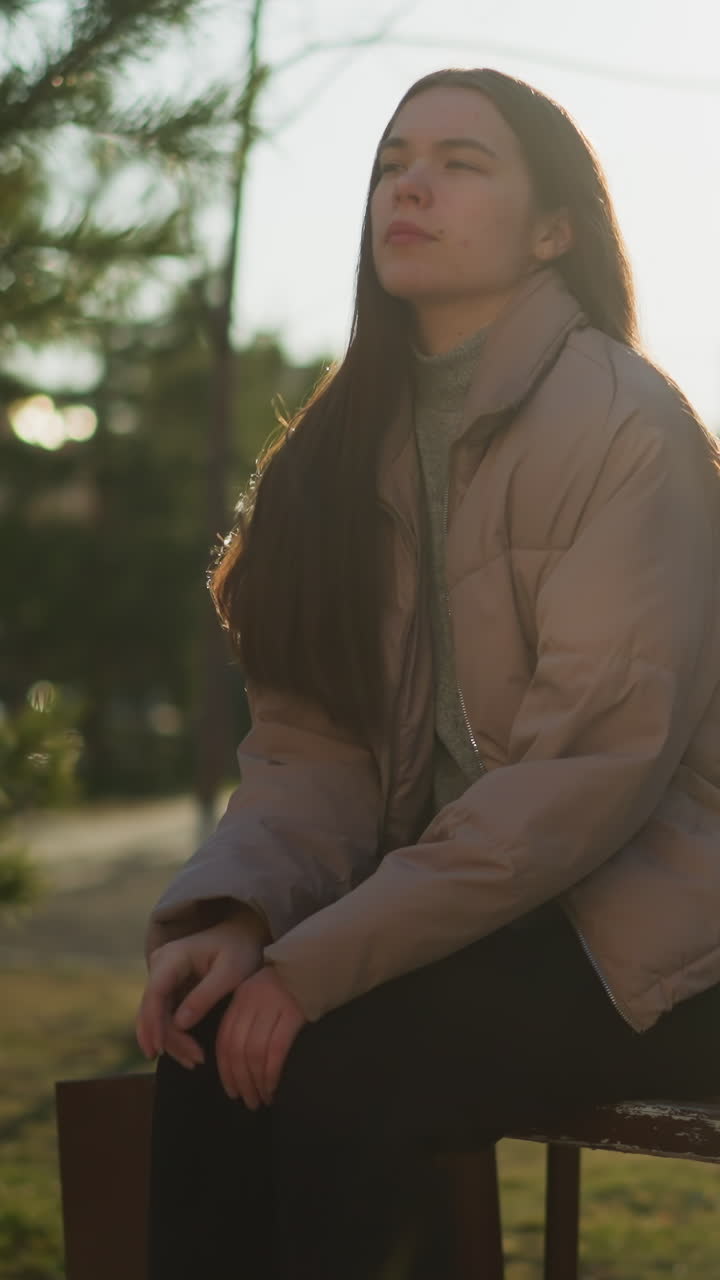 una joven con una chaqueta de melocotón, camisa interior gris y pantalones negros se sienta en un banco del parque con sus patines a su lado. descansa una mano en su barbilla en profundo pensamiento, con un fondo borroso
