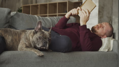 Mann,-Der-Mit-Seinem-Bulldoggenhund-Auf-Dem-Sofa-Liegt,-Während-Er-Ein-Buch-Liest-2