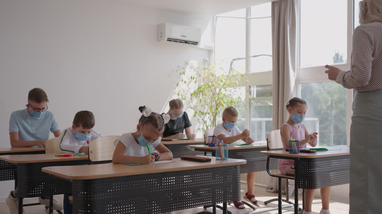 교실에서 선생님의 설명을 듣고 글을 쓰는 다민족 그룹의 아이들이 얼굴 마스크를 쓰고 수업에서 일하고 있습니다.