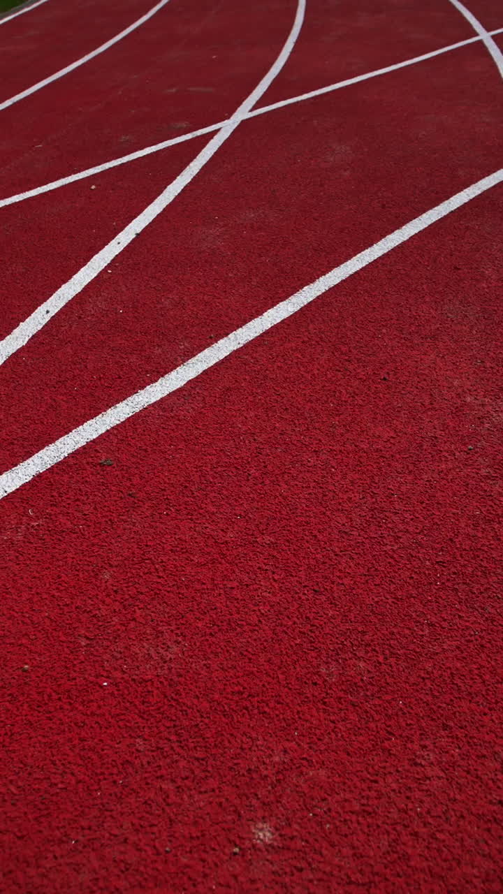 Following dark red running track crossed by the white lines. Green grass field and paved path on both sides of the track. Close up. Vertical video