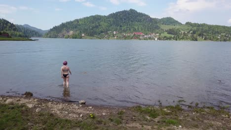 aerial approach shot, of girl with short purple hair walking into lake with black swimsuit on