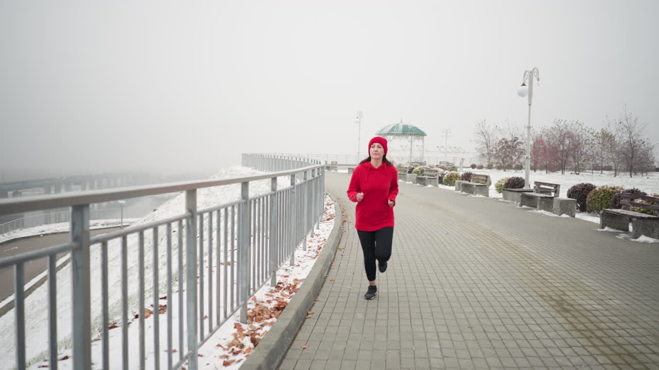 Athlete in sport attire jogging on interlocked pathway near iron railing in serene snowy park, surrounded by benches, foggy atmosphere, distant bridge, and vibrant bushes in winter scenery