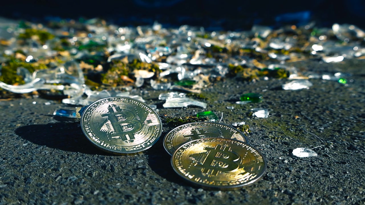 Monete fisiche di Bitcoin che giacciono sull'asfalto accanto a frammenti di vetro in frantumi, a simboleggiare i rischi, le perdite e la volatilità del mercato delle criptovalute, con la luce del sole che si riflette su di esse
