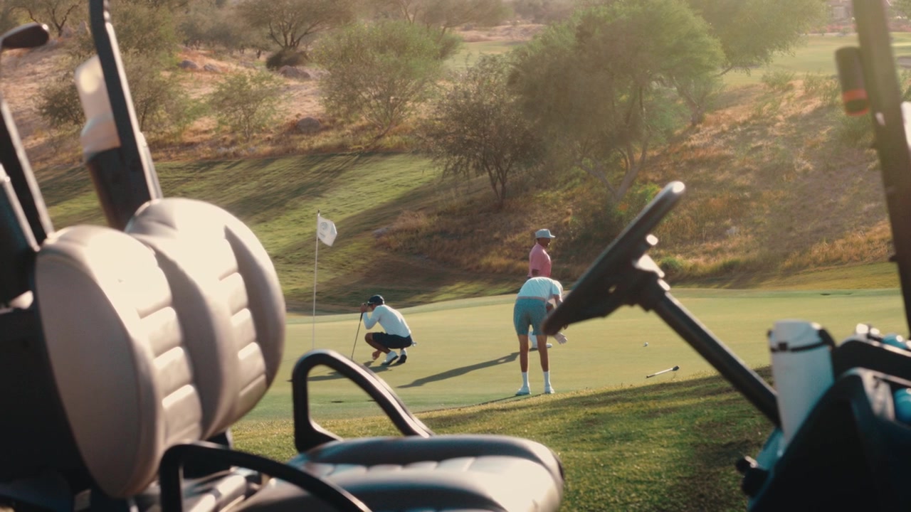 Two golfers read a putt on a desert green seen past a parked golf cart in warm golden-hour light, filmed in slow motion, great for golf lifestyle and resort promos.