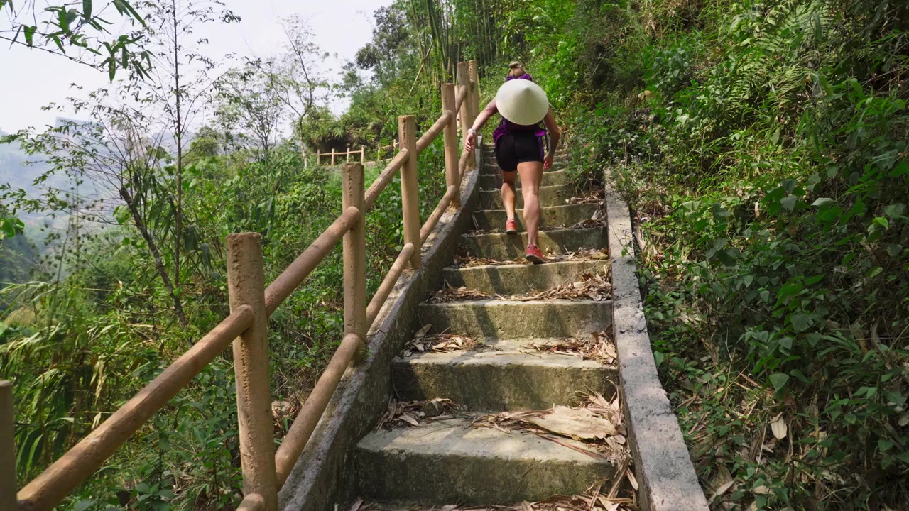 Một người đang leo cầu thang đá được bao quanh bởi cây xanh tươi tốt, đội nón lá truyền thống và đeo ba lô, tại Sa Pa, tỉnh Lào Cai, Việt Nam - Chuyển động chậm