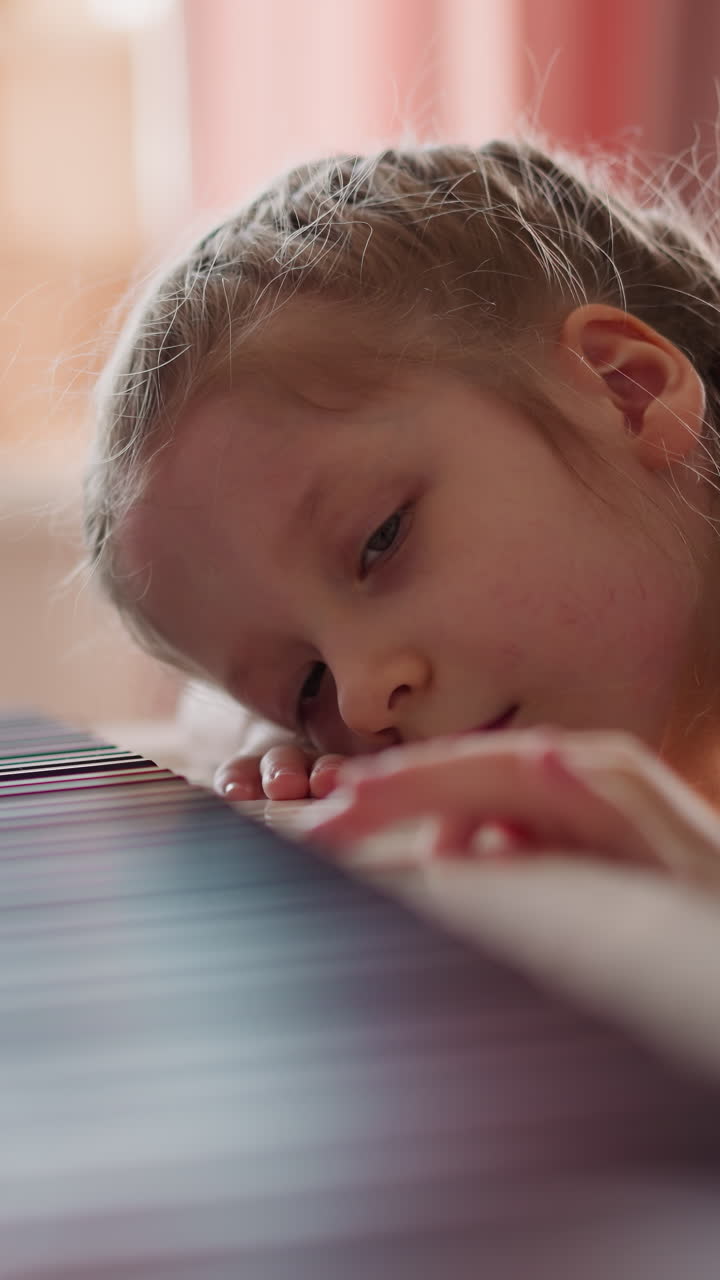 Tired junior schoolgirl with plaits presses piano keys leaning on instrument in living room. Little child bored of music lesson at home. Creative hobby