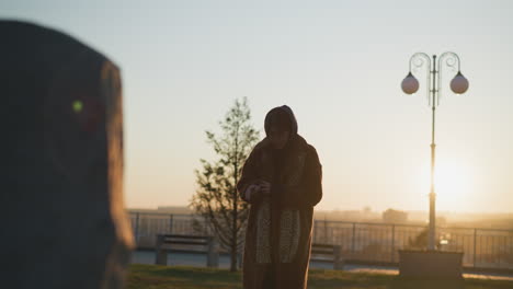 a tearful girl stands in front of a stone monument in a park , wearing a coat and scarf. her hands are folded in her arms, and she is in the process of removing her hood, revealing a sense of sorrow