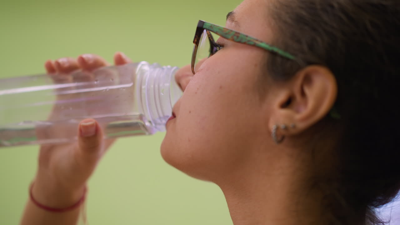 Close up view woman in white top and glasses drinking water indoors with calm relaxed face holding clear bottle soft natural light blurred studio background wellness recovery refreshing hydration