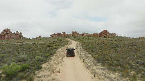 drone flies backwards away from rocks in desert landscape over a road where a black off-road vehicle is driving in the opposite direction in cederberg wilderness area in south africa