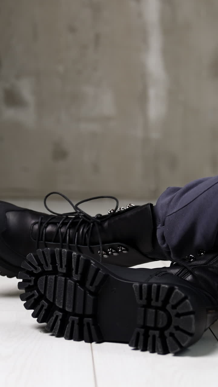Model in grey sportive pants sitting on the white floor in studio. Female touching the tongues of black stylish army boots on her feet. Vertical video