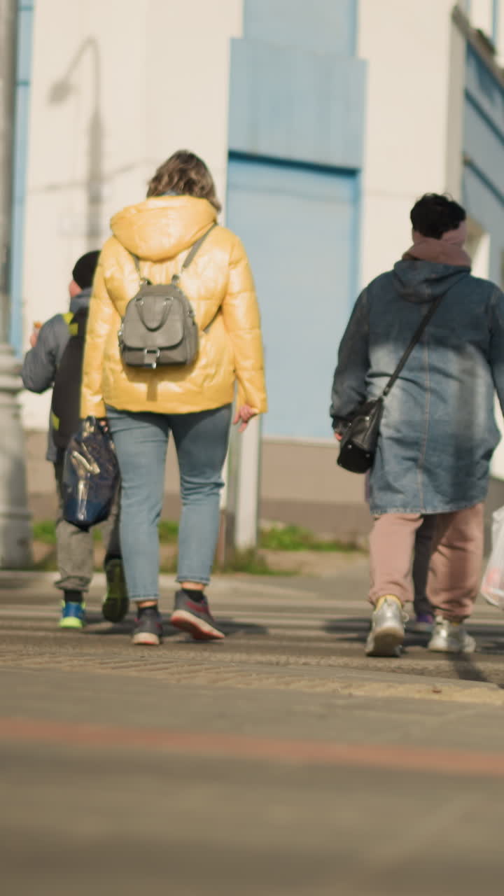 Madre blanca caminando con niños por la acera, chaqueta amarilla acolchada y mochila gris, llevando una bolsa de compra reutilizable, escolta protectora a través de un paso de peatones urbano, vaqueros informales y zapatillas