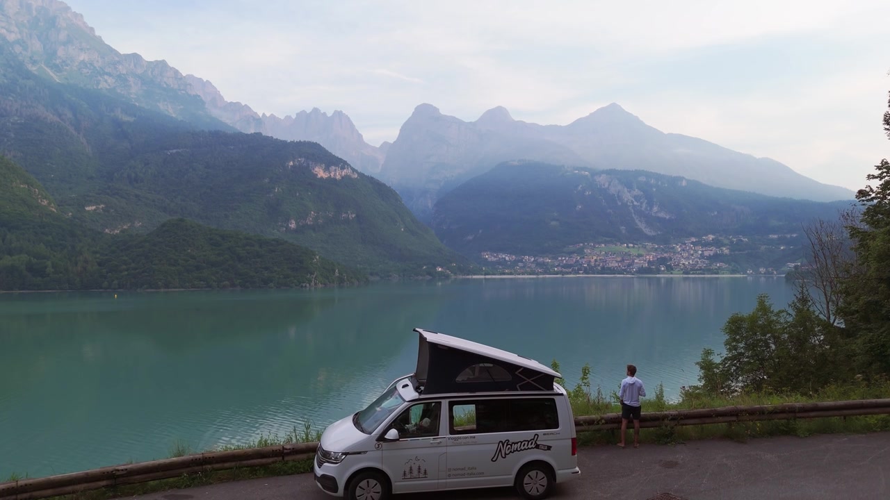 Un drone scende, riprendendo un viaggiatore solitario accanto a un camper, che sorseggia caffè e osserva il lago.
