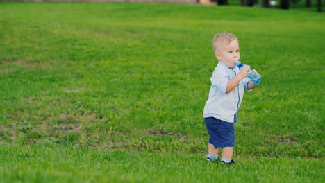 Funny-Kid-1-Year-Is-Drinking-Water-From-A-Bottle-It-Stands-On-A-Green-Lawn-Coolly-Staggers-Fun-Video