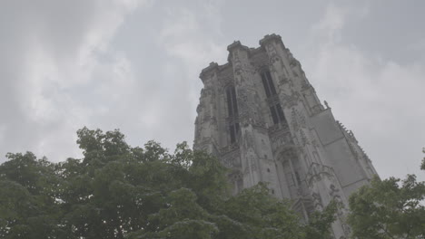 Rotierende-Tiefwinkelaufnahme-Des-Sint-Rombouts-Turms-In-Mechelen,-Belgien,-Mit-Einigen-Bäumen-Im-Vordergrund-An-Einem-Sonnigen-Tag-Mit-Blauem-Himmel-Und-Wolkenstamm