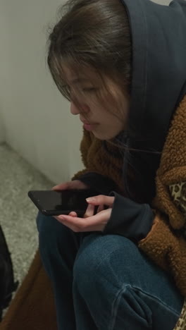 una chica con un abrigo marrón se sienta inclinada en un túnel subterráneo, sosteniendo su teléfono con una expresión de angustia. parece profundamente preocupada y perdida en sus pensamientos