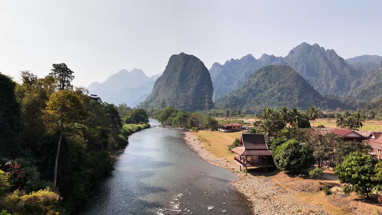 River winds through lush Pha Tang valley in Vang Vieng, Laos, flanked by forested mountains, traditional wooden houses, and tropical vegetation under clear blue sky in tranquil rural setting