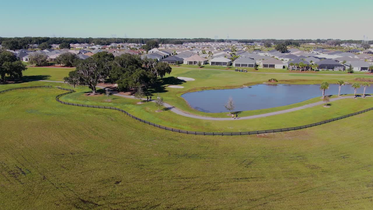Ancora un drone che mostra persone che camminano accanto a un laghetto fiancheggiato da palme, con un campo da golf nelle vicinanze e un complesso residenziale per pensionati che creano una scena di comunità all'aperto pacifica e attiva