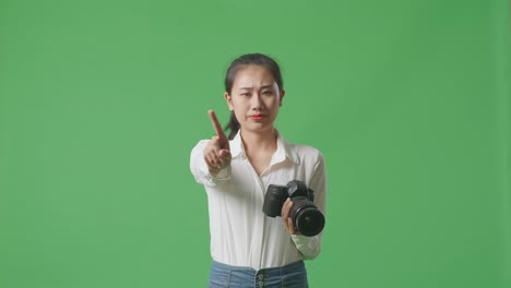 심각한 아시아 사진작가는 그녀의 손에 카메라를 들고 스튜디오에서 녹색 화면 배경에 서있는 동안 지표 손가락 표시 없이 불쾌감을 표합니다.