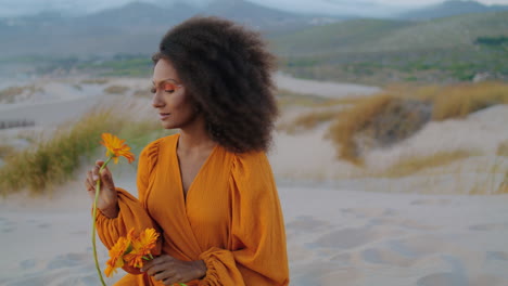 Portrait-relaxed-model-twilight-at-sandy-desert-sniffing-flower.-Woman-on-dunes.