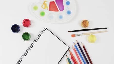 vista aérea del cuaderno de notas, pinturas, pinceles y lápices de colores sobre fondo blanco, en cámara lenta