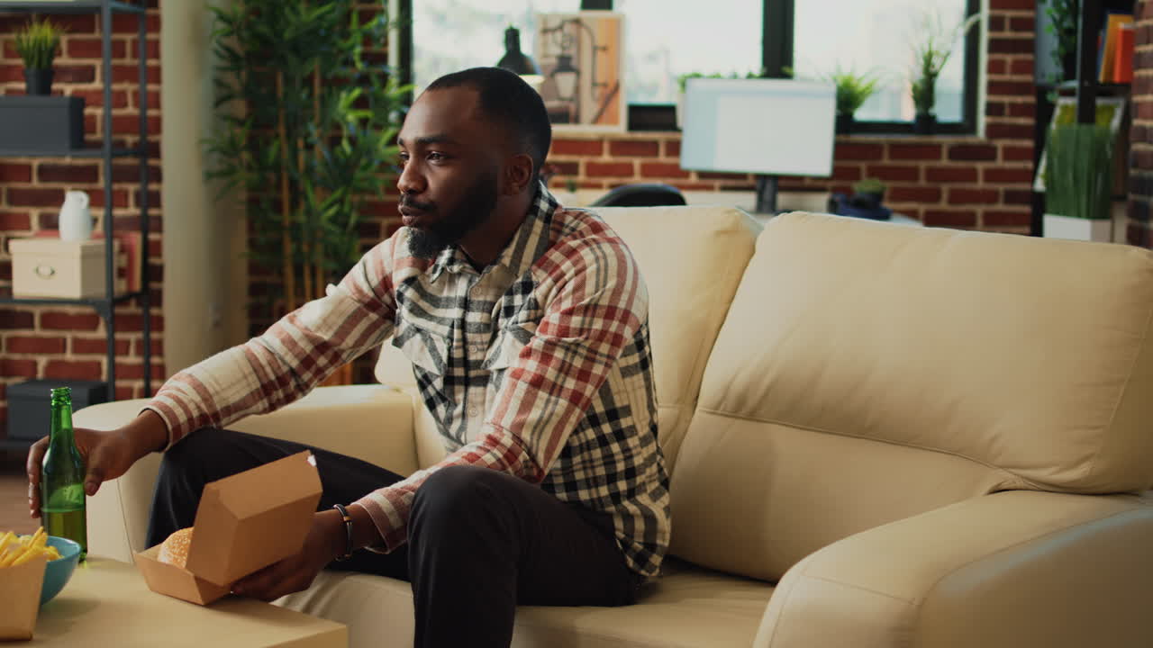 Modern man eating hamburger with fries on couch