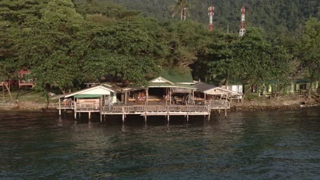 Drone-side-trucking-shot-of-empty-tourist-backpacker-style-restaurant-on-the-sea-with-telecommunication-towers-behind-on-Koh-Chang-Thailand,-due-to-the-effects-of-covid-on-travel-and-tourism