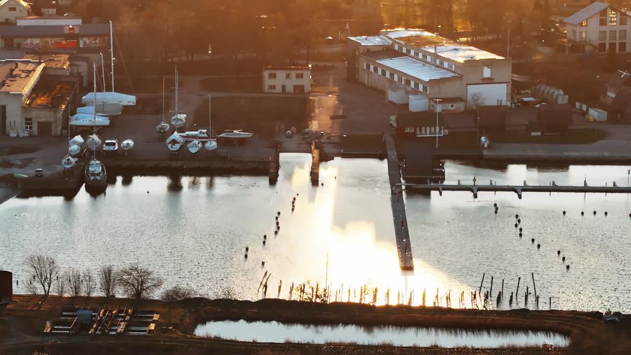 A warm golden glow highlights the empty docks, parked boats, and boatyard structures at Engure Port, as sunset bathes the harbor in serene evening light.