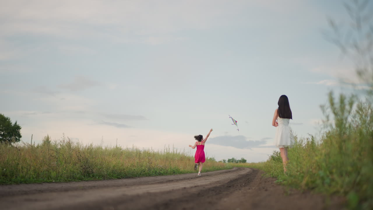 흑인 여성과 백인 여성이 황혼 무렵 시골 흙길에서 연을 날리며 장난기 넘치는 추격전과 응원의 웃음을 짓고, 다채로운 무지개 연이 하늘로 날아오르는 모습, 초원의 풀, 영화 같은 와이드 샷, 따뜻한 영화 톤