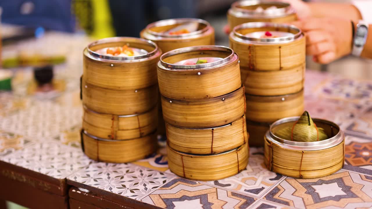 Bamboo steamers filled with dim sum on a patterned table, hands reaching in a vibrant market setting