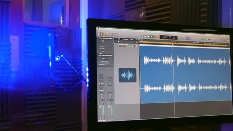 una forma de onda de audio en un monitor de computadora en un estudio de grabación con un micrófono visible en la cabina vocal detrás