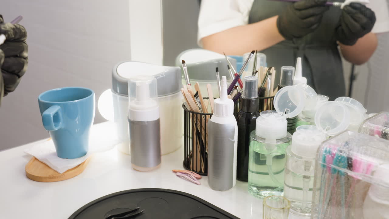 Closeup view of beautician in black gloves wetting cotton pad and cleaning eyebrow brush on white salon table with makeup tools, cotton rounds, solution bottles, and reflection in mirror