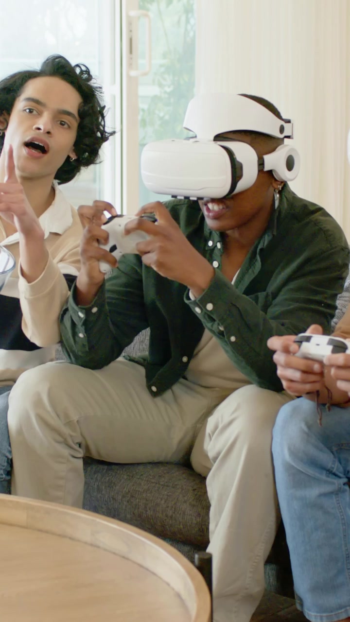 Video verticale: Uomo al centro che indossa un visore VR e gioca con un gamepad sul divano del soggiorno per un gioco di gruppo.