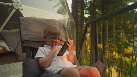 focused little boy watches movie on smartphone enjoying peace of evening nature. small boy relaxes alone propping cheek with finger feeling calm atmosphere