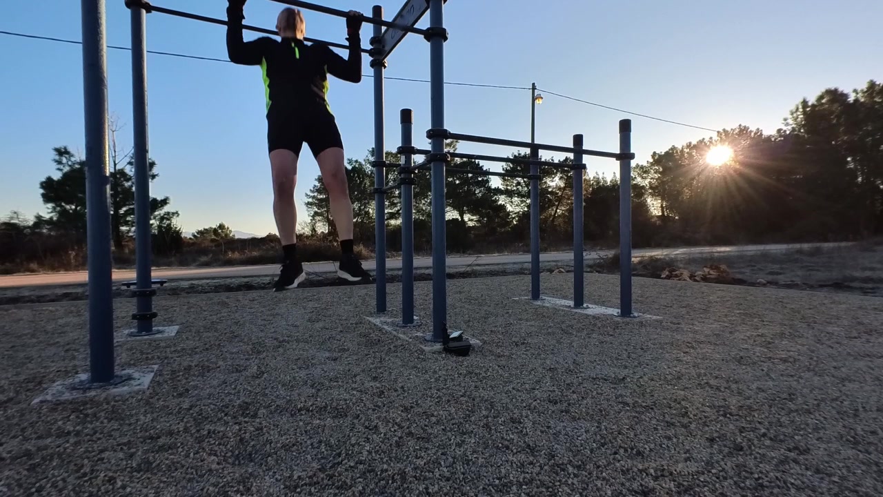 Front view of an athletic man doing pull-ups on a bar in an outdoor fitness area during sunrise. Natural backlight with trees in the background. Concept of bodyweight training and morning routine