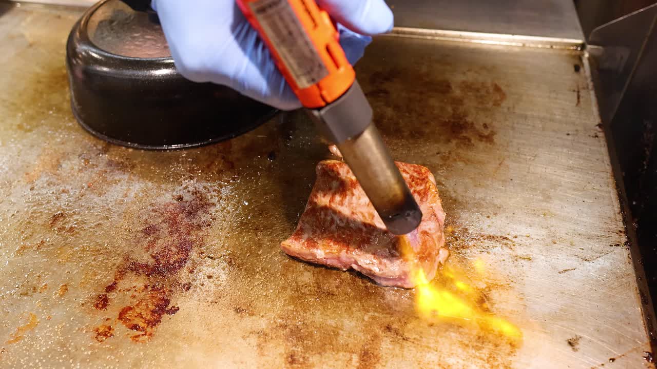 Primo piano di uno chef che sigilla manzo Wagyu marmorizzato usando una torcia da cucina su una griglia teppanyaki.