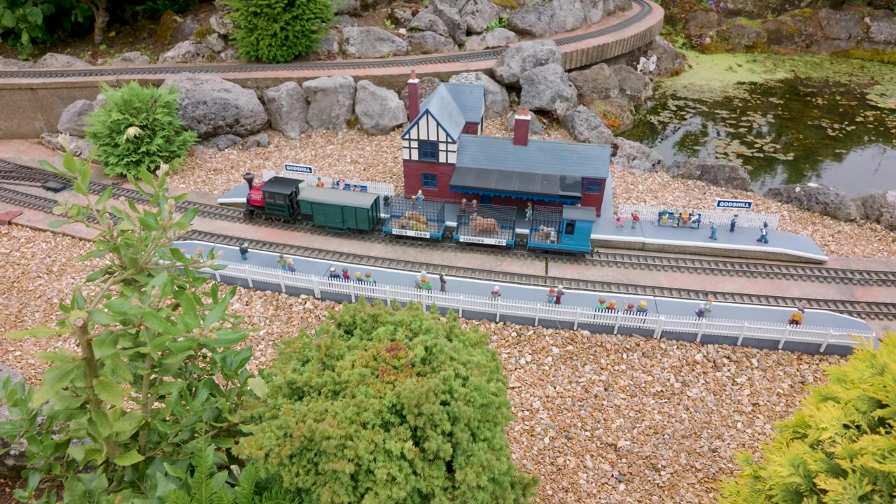 Trenini a vapore modello partono insieme alla stazione del villaggio modello di Godshill mentre locomotive in miniatura trainano carrozze oltre piccole figure, piattaforme ed edifici in un'esposizione dettagliata di ferrovia da giardino