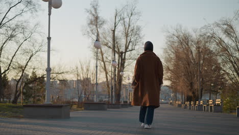 back shot of a girl walking somberly through a lonely park at sunset, wearing a brown coat and jeans. the scene captures the quiet solitude of the moment, with empty benches and bare trees