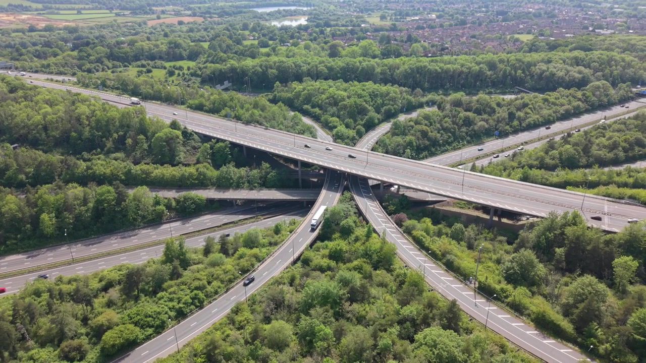 An aerial view captures vehicles moving along a complex highway interchange. Multiple lanes and bridges are surrounded by dense green trees under a bright daytime sky. Cars and trucks travel smoothly.